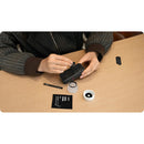 A person's hands are shown assembling a black smartphone camera lens protector kit on a light wood desk. The kit includes a black protective ring, a small black tool, a white lens cap, and a black instruction card at best camera store