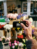 Hand holding a Fujifilm QuickSnap Black and White disposable camera against a background of colorful flowers at a market, offered by Best Camera Store.