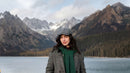 A woman wearing a gray tweed flat cap, green scarf, and gray coat stands in front of a scenic mountain and lake background.