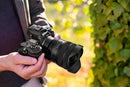A photographer holding a Nikon mirrorless camera equipped with the Tamron 12-20mm F2.8 Di III-A VXD lens up to his eye, showcasing outdoor action photography gear from Best Camera Store.