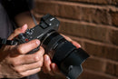 Hands holding the Tamron 16-50mm f/2.8 lens with lens hood attached to a Sony mirrorless camera from Best Camera Store.